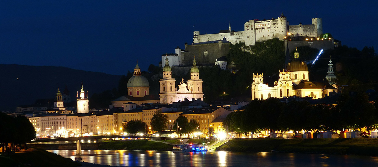 A large castle overlooks a city of multiple buildings nestled behind a river at night